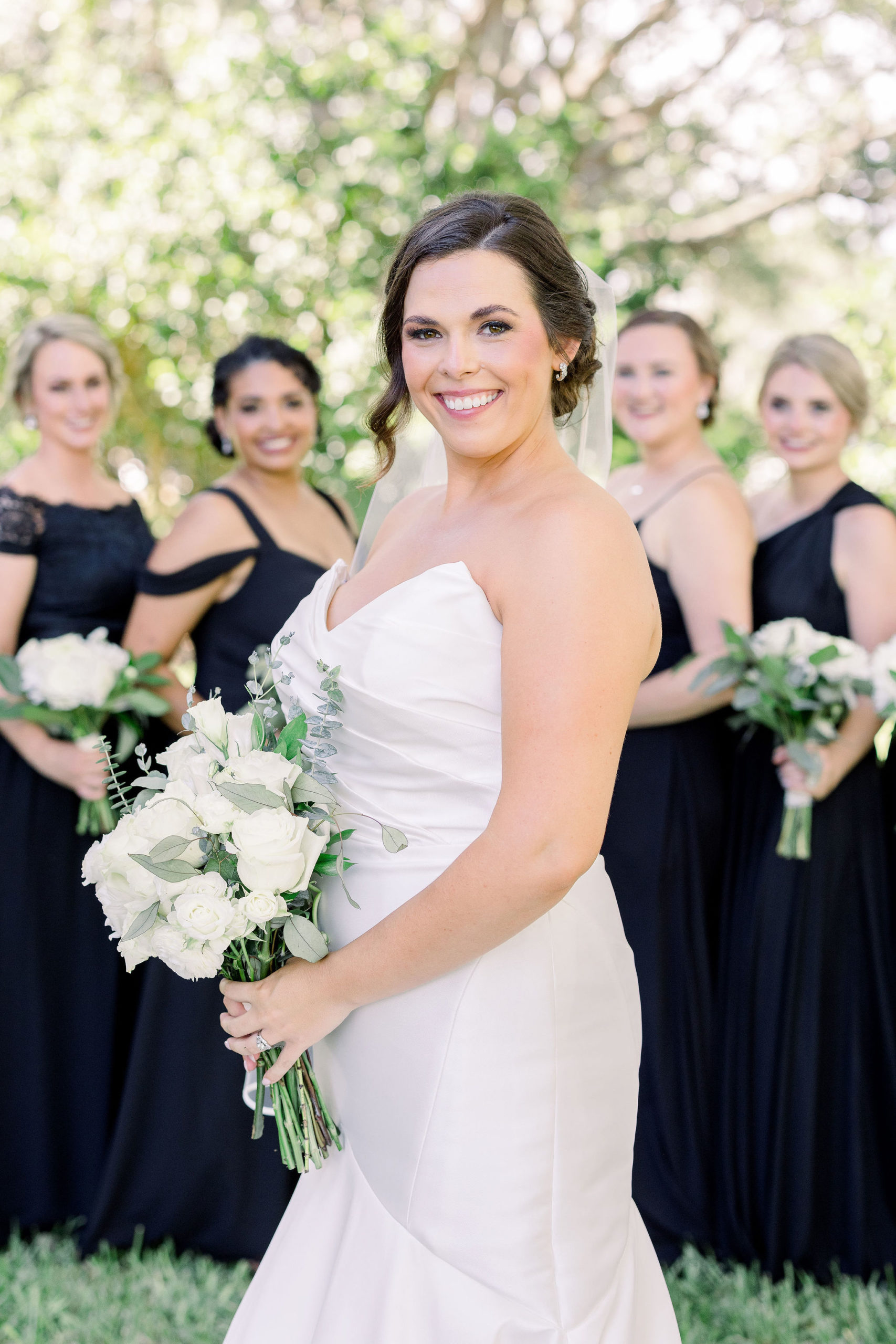 A bride wearing a mermaid strapless wedding gown with her bridesmaids in the background smiling on her wedding day by Jommy Photography. Black summer bridesmaid dresses with the bride in white for a timeless classic wedding style.
