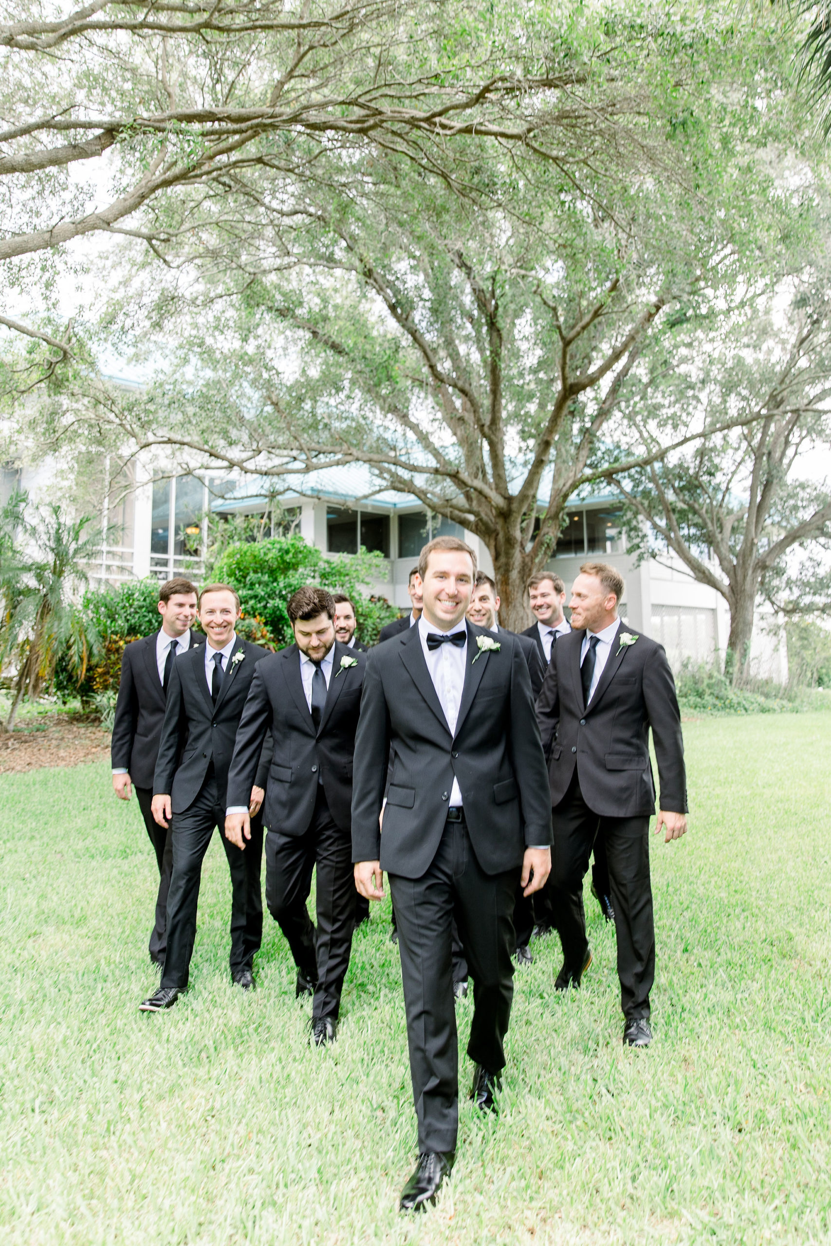 A groom walks with his group of groomsmen all wearing black suits and ties at the IMG Golf Course in Florida by Jommy Photography. A timeless groomsmen style ideas and inspiration for a summer wedding.
