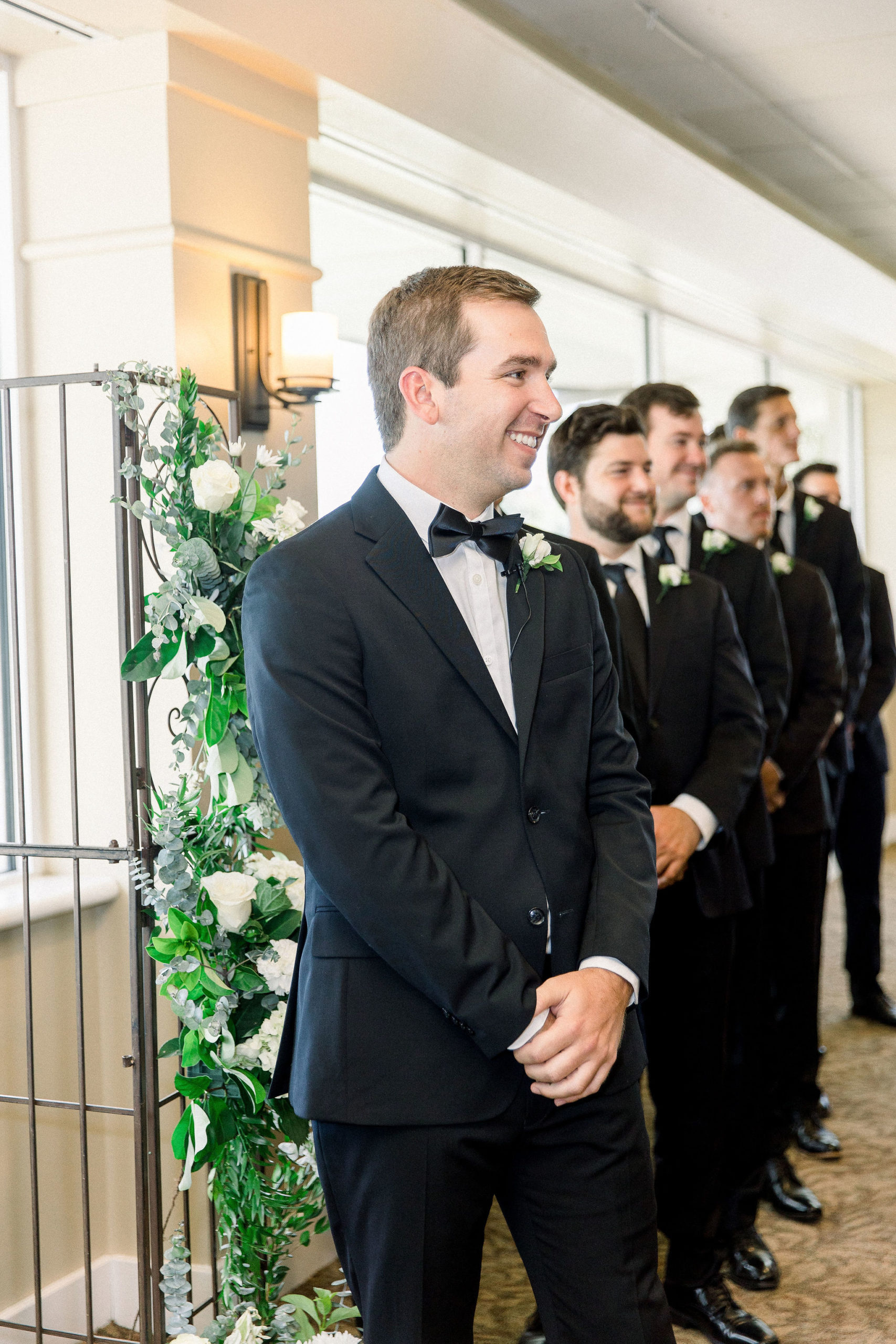 Groom in a black suit and a black bowtie stands with his groomsmen waiting for his bride with a smile on his face captured by Jommy Photography. Smiling groom waiting for his bride at the wedding altar.
