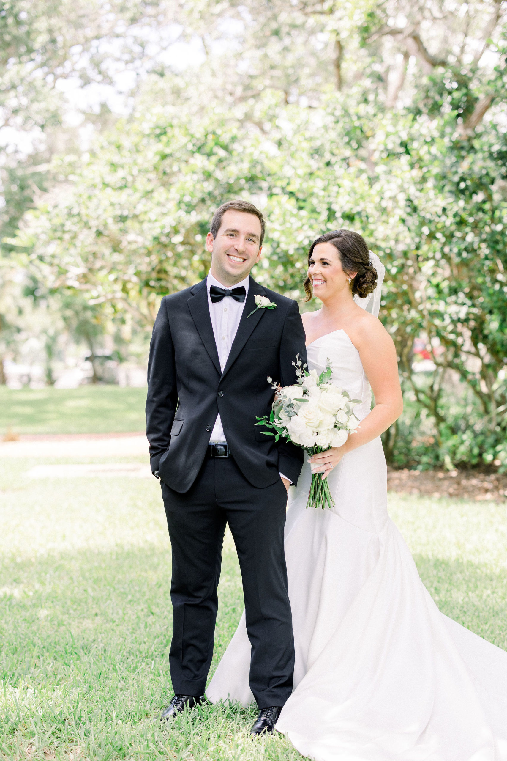 A bride with a simple white silk mermaid wedding gown smiles at her husband as he looks forward by Jommy Photography. Groom in a black bowtie and hands in his pocket smiles on his wedding day with his bride at the IMG Golf Course.
