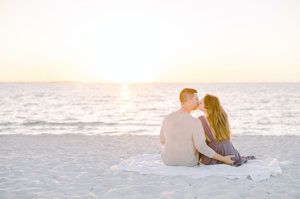 An engagement photographer in FL captures a couple sitting on the beach kissing by Jommy Photogrpahy. sunset #JommyPhotography #JommyEngagements #BeachEngagements #FloridaEngagements #FloridaBeachEngagement #ClearwaterFlorida #EngagementPhotographer
